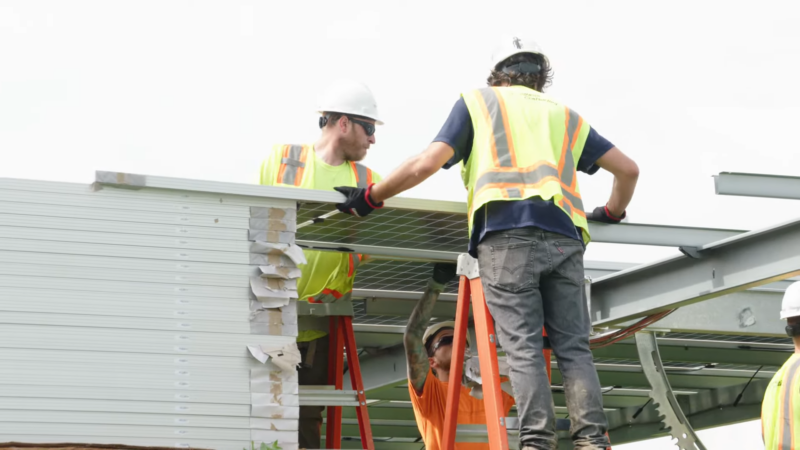 The Image Shows Construction Workers in Safety Gear, Including Helmets and Vests, Installing Solar Panels on A Metal Framework at A Solar Power Plant Site