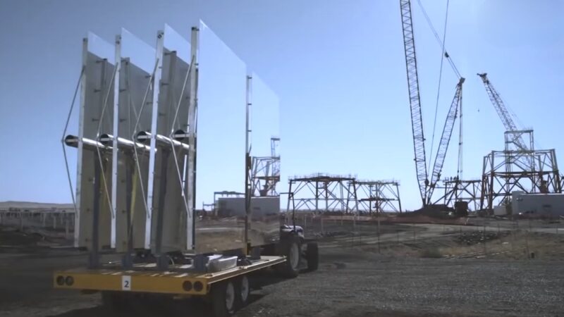 The Image Shows a Solar Power Plant Under Construction, Featuring Large Solar Panel Mirrors on A Mobile Platform with Cranes and Steel Frameworks in The Background