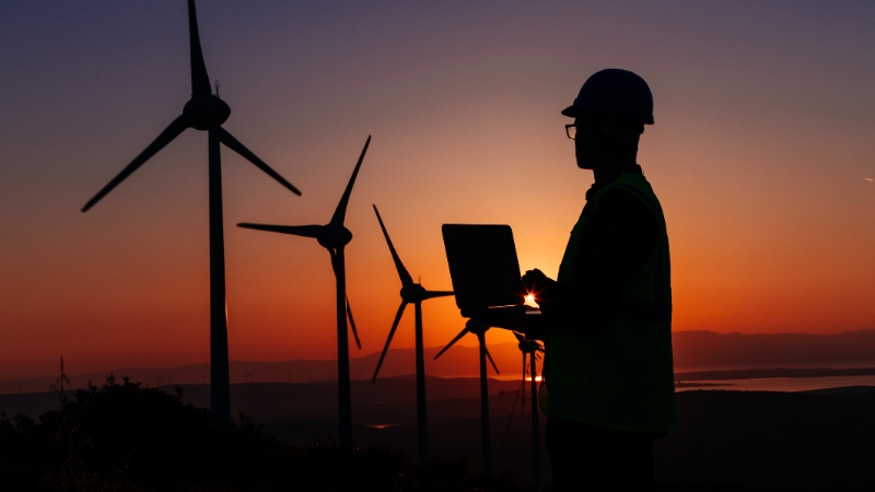 A silhouetted person in a hard hat looks at a laptop against a sunset backdrop with wind turbines