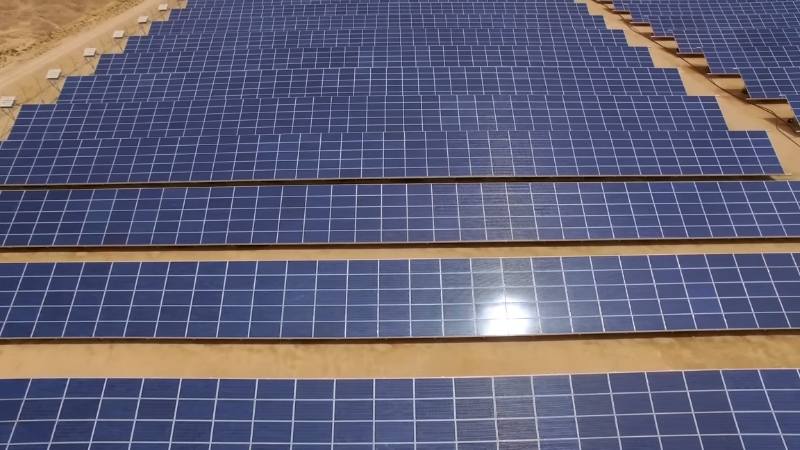 A vast array of blue solar panels stretches across a sandy, arid landscape under a bright sky