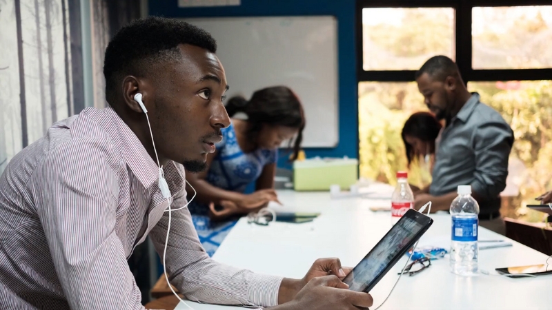 A young man uses a tablet with earphones in a shared workspace while other people work in the background on laptops and paperwork