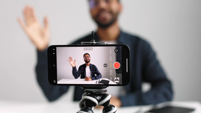 A man records himself waving on a smartphone mounted on a small tripod in a bright indoor setting