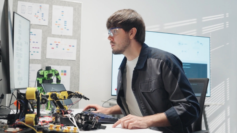 Engineer focuses on a computer screen while working with robotic components in a lab