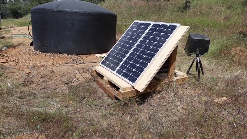 Small solar panel setup powering an off-grid irrigation system next to a water tank in a dry field