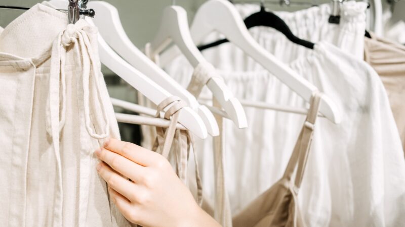 A hand checks neutral-toned slow fashion garments on a clothing rack